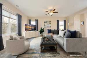 A virtually staged living room with beige walls, large windows, and a ceiling fan. The room features a gray sofa, a round armchair, a tufted ottoman, a TV on a stand, and a large rug. A potted plant and wall art add decorative touches.