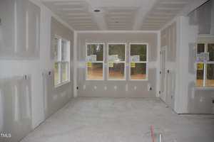 A room under construction with drywall installed and unfinished walls. There are several windows with stickers on them, a door, and an unfinished floor. The ceiling has recessed lighting fixtures.