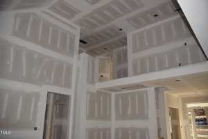 The image shows an interior view of a house under construction. The walls and ceiling are made of drywall panels, with visible joint tape and compound. Natural light enters through a window and a glass door on the right side.