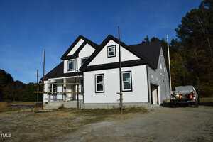 A partially constructed two-story house with white siding and black roof accents. Scaffolding is present on one side, and a truck with building materials is parked on the gravel driveway. Trees and clear skies are visible in the background.