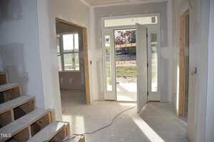 A partially completed interior of a house, featuring a white front door slightly open, exposing a view outside. Unfinished drywall and wooden staircase are visible, along with construction materials on the floor. Electrical wiring is seen on the walls.