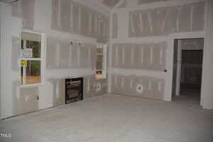 A room under construction with unpainted drywall on the walls and ceiling. There are two windows on one wall and a door frame on another. Electrical outlets are visible, and there is an uncovered floor.