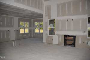 Interior view of a partially constructed room with unfinished drywall, exposed electrical outlets, and plumbing fixtures. There are large windows with a view of trees outside. A fireplace area is framed but uncompleted, and the floor is bare concrete.
