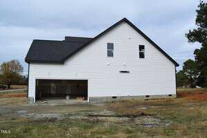 A partially constructed white house with a dark roof is shown. The garage lacks doors, and the windows have no frames. The surrounding area is grassy with some dirt patches and trees in the background.