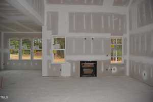 A room under construction with unfinished drywall on the walls and ceiling. There are several rectangular windows, and an opening for a fireplace in the center wall. The floor is made of bare concrete. The scenery outside the windows is wooded.