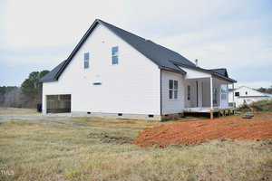 A newly constructed white house with a black roof sits on a grassy plot. It features a covered porch and a two-car garage. The surrounding area includes a patch of bare soil and a rural landscape with trees in the background.