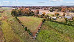 Aerial view of a triangular plot of grassy land enclosed by a fence. Surrounding the plot are fields with patches of green and brown. In the background, there are several trees and cloudy sky, along with a few buildings and roads.