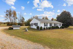 Located at 6206 Huntsboro Road, Oxford, this charming white single-story house features a gray roof and green shutters. It's nestled on a grassy lawn with shrubs lining the front. A gravel driveway guides you home, while a small shed rests peacefully under the clear blue sky with scattered clouds.