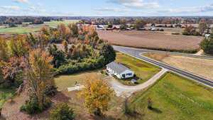Aerial view of a rural landscape featuring a single-story white house with a gray roof surrounded by trees. A paved road curves near the property, and fields stretch into the distance under a partly cloudy sky.
