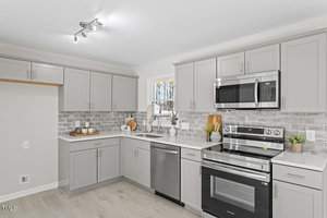 A modern kitchen with light gray cabinets, stainless steel appliances including an oven and microwave, and a tiled backsplash. The countertop features decorative items like wooden boards and a plant. The space is well-lit with ceiling spotlights.