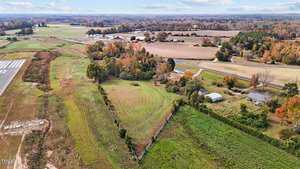 Aerial view of a rural area with fields and scattered trees. A triangular fenced pasture is in the foreground, and several homes and barns are visible. A small runway is on the left, and the landscape stretches into the horizon under a clear sky.