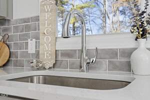 A modern kitchen sink with a shiny chrome faucet is set against a gray tiled backsplash. A wooden plaque with "WELCOME" hangs beside, and a vase with decorative branches sits on the countertop. A window reveals a view of trees outside.