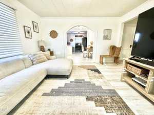 A cozy living room with a light-colored sectional sofa on a patterned rug. There is a large flat-screen TV on a wooden stand. The space opens to a kitchen through an arched doorway. White walls and light wood floors enhance the bright, airy feel.