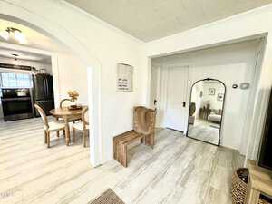 A bright room with light-colored flooring and walls. A wooden bench and two wicker baskets are near a large mirror. Adjacent is a dining area with a round table and chairs, leading to a kitchen with dark appliances. A sign on the wall reads about love.
