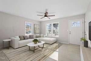 A modern living room with white walls and light wood flooring. It features a large L-shaped sofa, a round coffee table with plants, a ceiling fan, and a rug. Three windows overlook a wooded area, and a door is next to the windows. A wall-mounted TV is on the right.