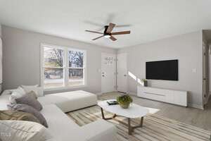 A modern living room with a white sectional sofa, gray and purple cushions, a round coffee table with a plant, wall-mounted TV, and a beige striped rug. The room features a ceiling fan and large windows with a view of a tree-lined street.