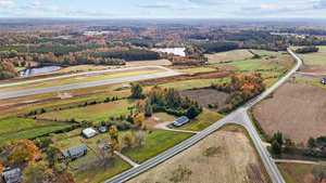 Aerial view of a rural landscape featuring an airstrip, fields, and a few houses. Roads intersect in the foreground. Trees, some with autumn foliage, and a distant lake can be seen under a partly cloudy sky.
