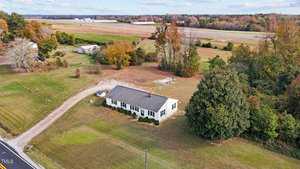 A small white house with a dark roof is surrounded by grass and trees in an open countryside setting. A dirt driveway curves from the road to the house. The background features fields, trees in autumn colors, and a clear sky.