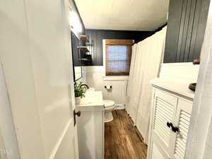 A small bathroom with wood flooring, white walls, and dark wall paneling. It features a white vanity with a mirror, a toilet, a shower with a beige curtain, a window with a bamboo shade, and a white cabinet with louvered doors.