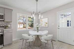 A modern dining area with a round white table and four light gray chairs on a patterned rug. A pendant light hangs above the table. A window and a door let in natural light. The walls are light-colored, and there's a framed artwork on one wall.