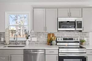 Modern kitchen with light gray cabinets and stainless steel appliances, including a stove and microwave. A tiled backsplash in shades of gray and white complements the countertop. A window above the sink displays outdoor trees. Decor includes a welcome sign.