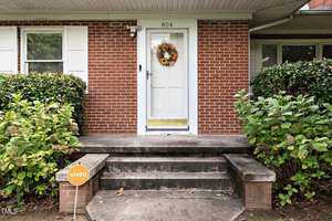 A white front door with a sunflower wreath is centered on a brick facade, flanked by green shrubs and white shutters. Three concrete steps lead up to the porch. A small Vivint security sign is staked in the grass to the left.
