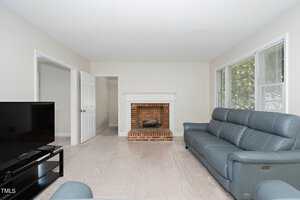 A living room with light gray walls featuring a brick fireplace and a white mantle. There are two blue leather sofas facing a large flat-screen TV on a black stand. The floor is covered with a transparent protective sheet. A window on the right lets in natural light.
