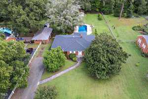 Aerial view of a single-story brick house at 604 18th Street, Butner, featuring a dark roof and surrounded by trees and a lush lawn. A swimming pool graces the backyard. The driveway elegantly curves toward the house, with a charming smaller building nearby in this verdant haven.