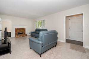 A living room with white walls and carpeted floor features a brick fireplace, two blue sofas, a black coffee table, and a television. A large window lets in natural light. The room leads to an entryway with a white door.