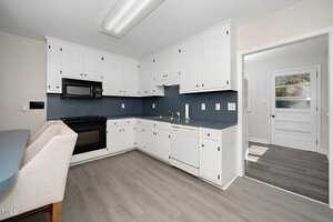 A kitchen with white cabinetry, a black microwave, and an oven. The countertops are blue, and there is a dishwasher. The floor is light gray wood, and there is a view of an adjacent room and a door leading outside.