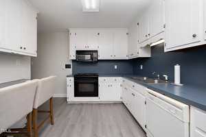 A kitchen with white cabinets, blue countertops, and a blue backsplash. It features a black oven with a stove, a microwave, and a dishwasher. Two beige stools are at a counter. The floor is light gray wood.