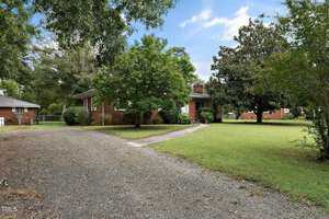 A brick house is surrounded by trees and a grassy lawn. A gravel driveway curves to the entrance, and another house is visible in the background. The sky is partly cloudy.