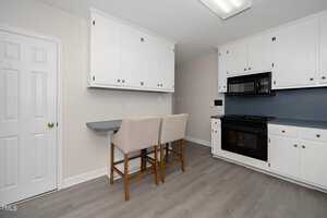 A bright kitchen features white cabinets, a black built-in oven and stove, and a blue backsplash. Two beige upholstered chairs sit at a small counter area. The kitchen has light wood flooring and a white door on the left.