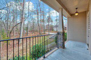 A porch with a tiled floor and a black metal railing overlooks a wooded area. The sky is clear with some clouds. The porch features a stone pillar and a hanging light. A path leads through the trees, lined with bare branches and sparse leaves.