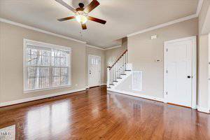 A bright living room with hardwood floors and neutral-colored walls. A ceiling fan hangs from the center of the white ceiling. There's a large window, a front door with glass panels, and a staircase leading to the upper floor.