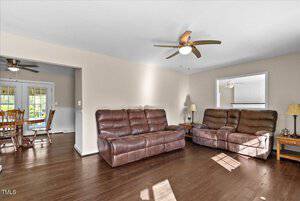 A spacious living room features two brown leather sofas and wooden side tables with lamps. The room has hardwood flooring and a ceiling fan. An adjacent dining area with a ceiling fan is visible through a wide doorway. Natural light enters through windows.