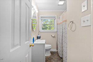 A small bathroom features a sink with toiletries, a circular mirror, and a toilet. The shower has a patterned curtain. There are two windows with blinds, and the walls are painted in a light color. A towel ring is attached to the wall.