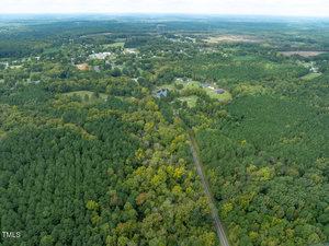 Aerial view of a lush, green forested landscape intersected by a narrow, winding road. In the background, a small town with dispersed buildings is visible. The sky is overcast, giving the scene a muted lighting.