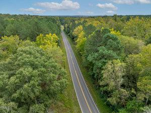 Aerial view of a straight, two-lane road surrounded by dense green foliage and trees. The road extends into the distance, splitting the lush forested area. A clear blue sky with scattered clouds is visible above.