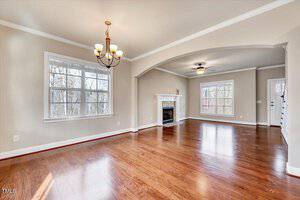 A spacious living room with polished wooden floors and beige walls. A chandelier hangs from the ceiling. There is a fireplace with a mantel, two large windows, and a ceiling fan. Natural light enters through the windows, offering a view of trees.