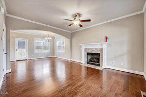 An empty living room with hardwood floors, cream-colored walls, and a ceiling fan with a light. A fireplace with a stone surround is on one wall. Large windows let in natural light. An archway leads to another room, and a door opens to the outside.