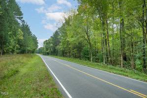 A rural two-lane road stretches into the distance, bordered by lush green trees on both sides. The sky is partly cloudy, and the road is flanked by a narrow grass verge. The scene is tranquil and serene.