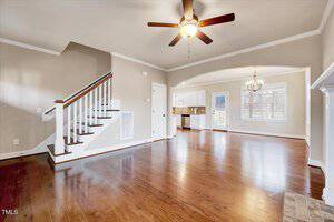 An empty living and dining area with polished hardwood floors and beige walls. The room has a ceiling fan, staircase on the left, and an archway leading to a kitchen with white cabinets. Natural light streams through a large window.
