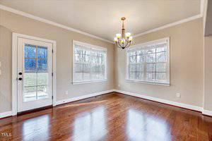 A bright room with polished hardwood floors, two large windows, a glass-paneled door, and a simple chandelier hanging from the ceiling. The walls are painted light beige, and there is a view of trees through the windows and door.