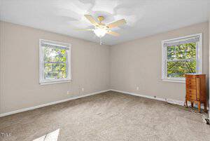 An empty room with beige walls and carpeted floor. Two windows with white frames allow natural light, and a ceiling fan is centered above. A small wooden dresser stands in the corner.