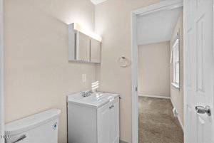 A small bathroom features a white vanity with an integrated sink and a wall-mounted mirror cabinet above. There is a toilet to the left. A doorway leads to a carpeted room with a window, visible in the background.
