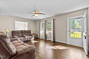 A spacious living room with hardwood floors, beige walls, and a ceiling fan. It features brown leather reclining sofas, two lamps, and large windows with curtains. An open door reveals a view of a green lawn and trees outside.