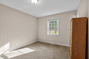 A small, empty room with beige walls and carpeted flooring. A wooden dresser stands in the corner near a window that lets in natural light, illuminating the room. A ceiling light fixture is centered above.