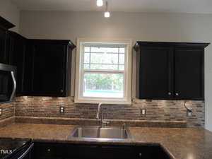 A kitchen interior featuring a central window above a stainless steel sink with a single faucet. The backsplash has a mosaic tile design. Dark cabinets with metal handles frame the sink area. A microwave is visible on the left. The countertop is laminate.