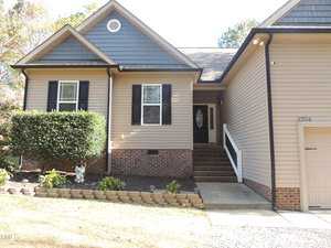 A tan house with black shutters and brick foundation. There is a small staircase leading to a front door with a glass oval window. A bush and some small plants are next to the steps. The house number is visible on the siding near the entrance.